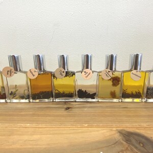 May include: Ten small glass bottles with silver caps and labels. The labels are round and have different names written on them, including "Amber", "Tiger", "Vanilla", and "Chamomile".