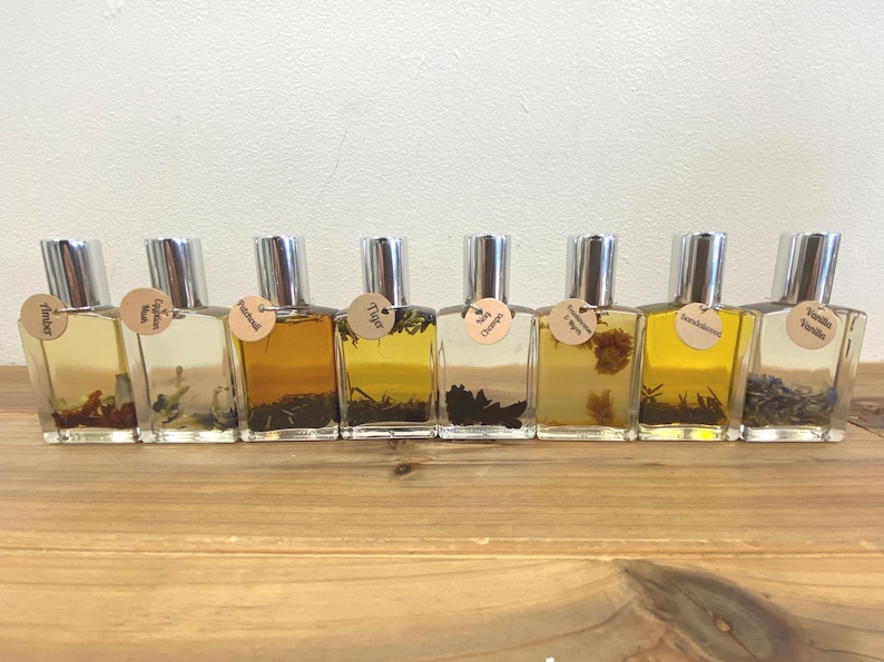May include: Ten small glass bottles with silver caps and labels. The labels are round and have different names written on them, including "Amber", "Tiger", "Vanilla", and "Chamomile".