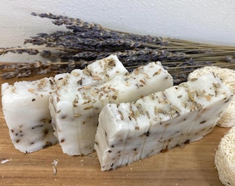 Peut inclure: Quatre savons faits à la main avec une base blanche et des flocons de lavande et d'herbes séchées. Le savon est disposé sur une surface en bois avec des tiges de lavande séchées en arrière-plan.