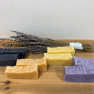 May include: A set of six handmade soap bars. Two bars are black, two are yellow, and two are purple. The soap bars are arranged on a wooden surface with a bundle of lavender.