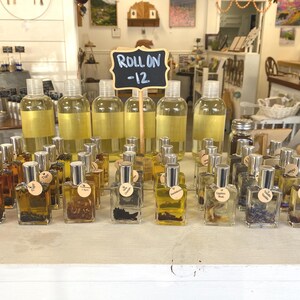 May include: A display of small glass bottles filled with various liquids and dried flowers. The bottles have silver caps and are arranged in rows on a white surface. A sign above the bottles reads "Roll On -12".