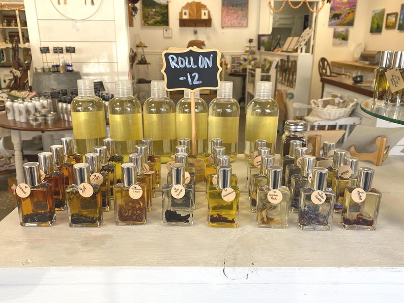 May include: A display of small glass bottles filled with various liquids and dried flowers. The bottles have silver caps and are arranged in rows on a white surface. A sign above the bottles reads "Roll On -12".