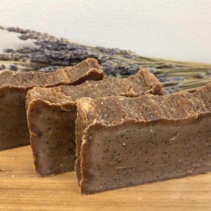 May include: Three bars of brown soap with a speckled texture, stacked on top of each other. The soap is sitting on a wooden surface with a bundle of dried lavender flowers in the background.