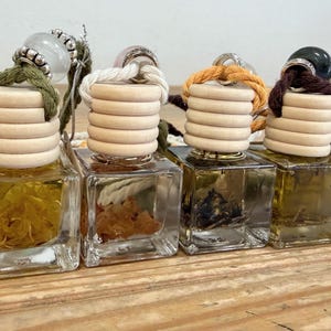 Könnte beinhalten: Eine Reihe quadratischer Glasflaschen, gefüllt mit gelber Flüssigkeit und botanischen Elementen. Jede Flasche hat einen gerippten Holzverschluss, eine dicke Aufhängeschnur in verschiedenen Farben und eine dekorative Edelsteinperle.