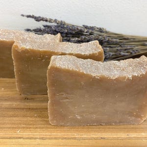 May include: Three bars of handmade soap, each with a light brown colour and a rough texture. The soap is arranged in a row on a wooden surface.