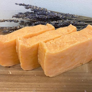 May include: Three bars of orange-colored soap, stacked on top of each other, on a wooden surface. The soap has a textured appearance.