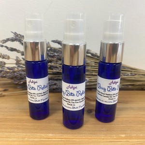 Puede incluir: Tres botellas de vidrio azul con tapas plateadas y etiquetas blancas. Las etiquetas dicen "Bug Bite Relief" y enumeran los ingredientes. Las botellas están sobre una superficie de madera con flores de lavanda secas.