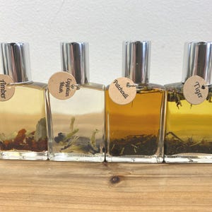 May include: Four small glass bottles with silver caps and brown labels. The labels have the words "Amber", "Egyptian Musk", "Patchouli", and "Tiger" printed on them. The bottles are filled with a clear liquid and contain small pieces of dried herbs.