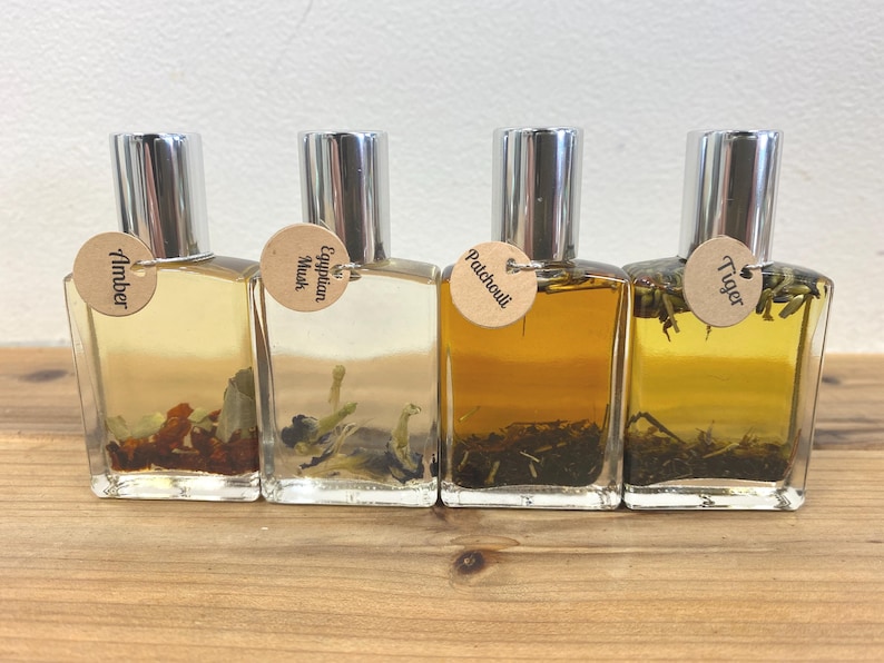 May include: Four small glass bottles with silver caps and brown labels. The labels have the words "Amber", "Egyptian Musk", "Patchouli", and "Tiger" printed on them. The bottles are filled with a clear liquid and contain small pieces of dried herbs.