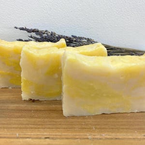 May include: Three bars of handmade soap with a yellow and white swirl pattern. The soap is arranged in a row on a wooden surface.