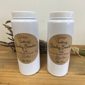 May include: Two white plastic bottles of Ashjoi Soothing Body Powder with Vanilla and Lavender. Each bottle has a brown label with text. Dried lavender and a bundle of herbs are behind the bottles. The bottles are on a wooden surface.