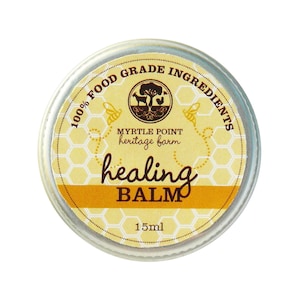Puede incluir: Una pequeña lata de plata con una etiqueta amarilla que dice "100% Food Grade Ingredients", "Myrtle Point Heritage Farm", "healing BALM" y "15ml". La etiqueta tiene un patrón de panal y una pequeña imagen de una granja con una cabra y una gallina.