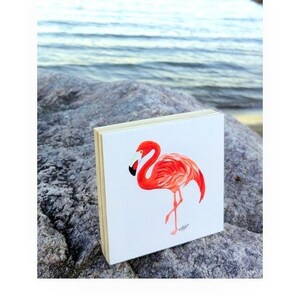 May include: A pink flamingo painted on a small wooden block. The flamingo is standing on one leg and has its head turned to the side. The background is white.