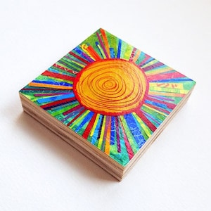 May include: A colorful wooden square with a painted sun design. The sun is yellow with red lines and is surrounded by colorful lines radiating outward.