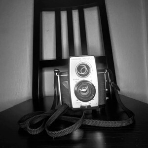 May include: A black and white Kodak Brownie Starflash camera with a black strap. The camera is sitting on a wooden surface.