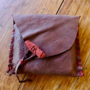 Può includere: Una borsa in pelle marrone con una patta in pelle rossa e un cinturino in pelle rossa. La borsa è chiusa e ha una cucitura decorativa attorno ai bordi.