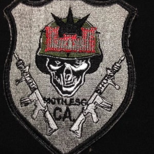 Puede incluir: Un parche bordado gris y plateado con una calavera que lleva un casco y una corona. La calavera tiene un diseño negro y rojo en el casco. Dos rifles cruzados están detrás de la calavera. El texto "South Central CA" está debajo de la calavera.