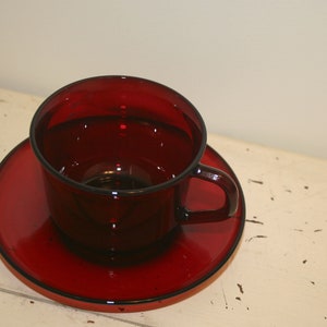 Può includere: Un set di tazza e piattino da tè in vetro rosso. La tazza e il piattino sono entrambi di un colore rosso intenso e ricco.