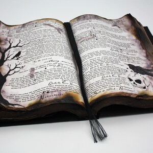 May include: An open antique book with a black and white illustration of a tree with a crow on a branch on the left page. The right page has a black and white illustration of a crow perched on a skull. The book has a black tassel.