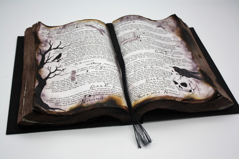 May include: An open antique book with a black and white illustration of a tree with a crow on a branch on the left page. The right page has a black and white illustration of a crow perched on a skull. The book has a black tassel.