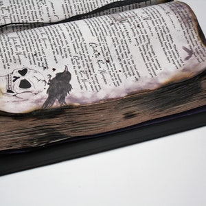 May include: A vintage book with a black cover and a skull and raven illustration on the front page. The pages are aged and have a burnt look. The book is open to a page with handwritten text.