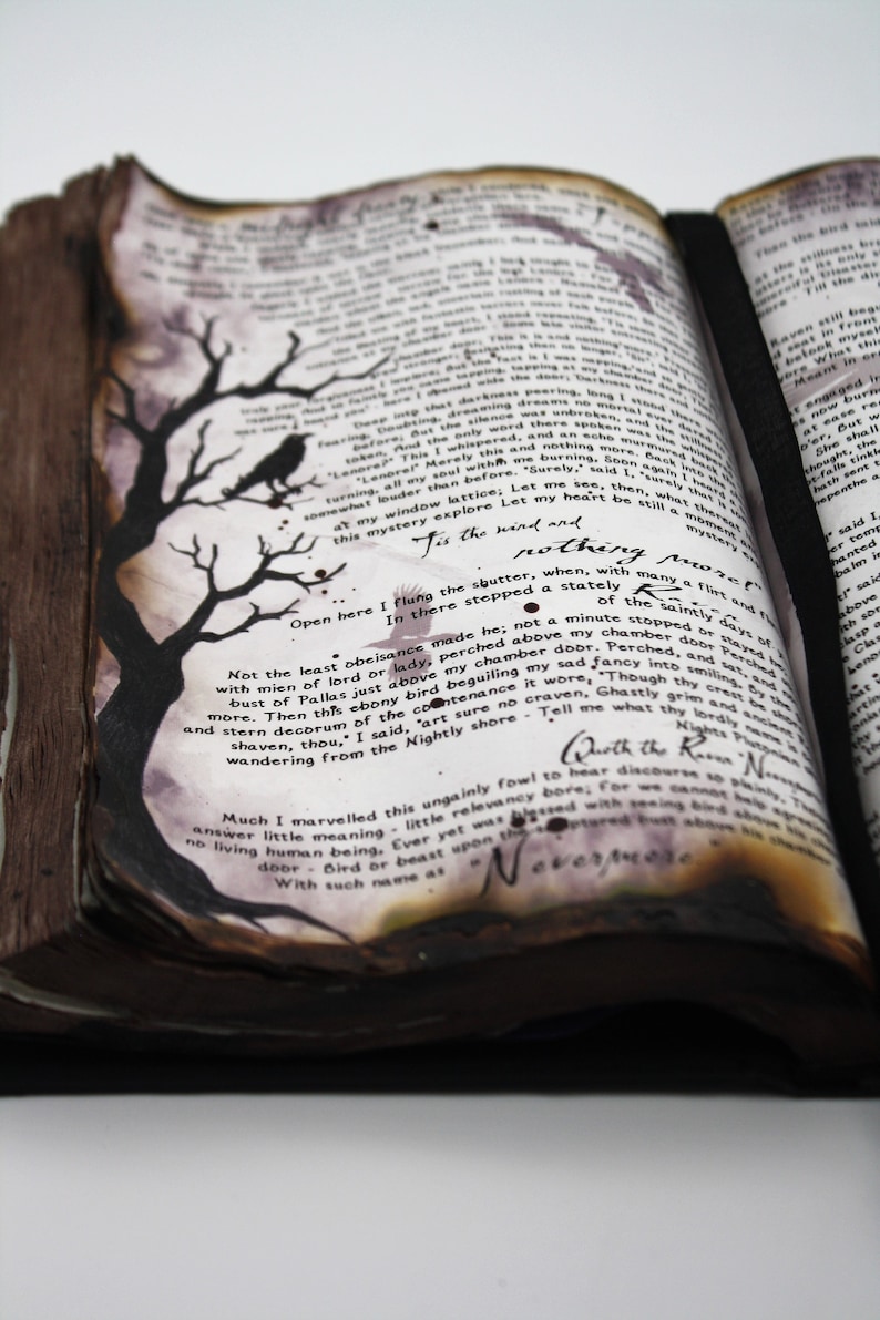 May include: An open book with aged, yellowed pages and a black leather spine. The pages are filled with handwritten text and illustrations of a raven and a tree. The text includes the phrase "Tis the wind and nothing more."