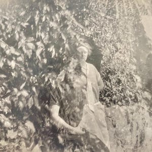 May include: A vintage black and white photograph of a person standing near foliage and a rock formation. The person is wearing a light-colored garment. The image has a soft focus and a nostalgic feel.