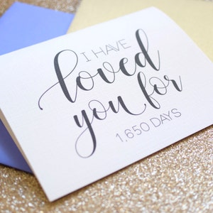 May include: White card with black calligraphy that reads "I have loved you for 1,650 days". The card is sitting on a gold glitter background.