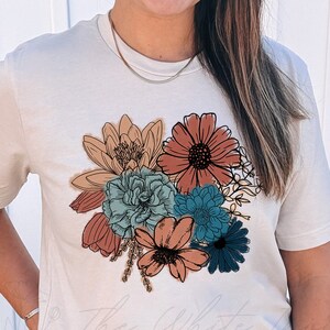 May include: A white t-shirt featuring a floral design with a variety of flowers in shades of orange, red, blue, and green. The flowers are outlined in black and have a vintage style.