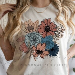 May include: A white t-shirt featuring a floral design with pink, peach, blue, and green flowers. The text "Personalized Gifts" is printed on the bottom of the shirt.