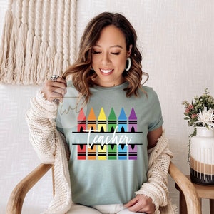 May include: A teal blue t-shirt with a rainbow of crayons and the word "Teacher" in a split monogram style.