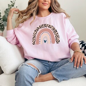 May include: A pink sweatshirt with a rainbow design featuring a leopard print pattern. The rainbow has a heart in the centre and is in shades of pink, beige, and grey.