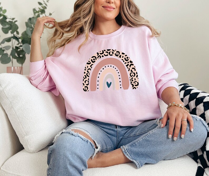May include: A pink sweatshirt with a rainbow design featuring a leopard print pattern. The rainbow has a heart in the centre and is in shades of pink, beige, and grey.