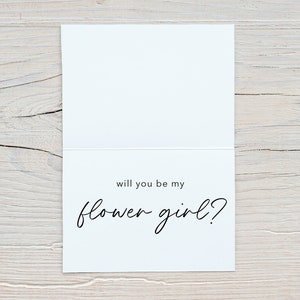 May include: A white card with black text that reads "Will you be my flower girl?"