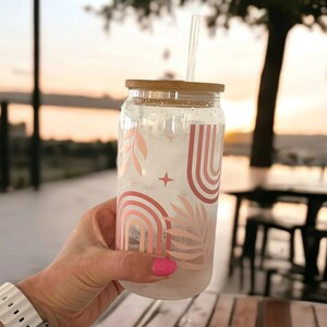 May include: A clear glass tumbler with a bamboo lid and a clear straw. The tumbler is decorated with a pink and peach rainbow and leaf design. The tumbler is filled with ice and held by a person.