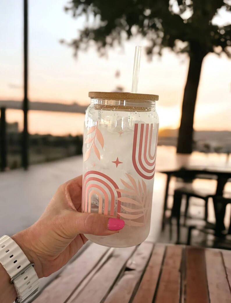 May include: A clear glass tumbler with a bamboo lid and a clear straw. The tumbler is decorated with a pink and peach rainbow and leaf design. The tumbler is filled with ice and held by a person.