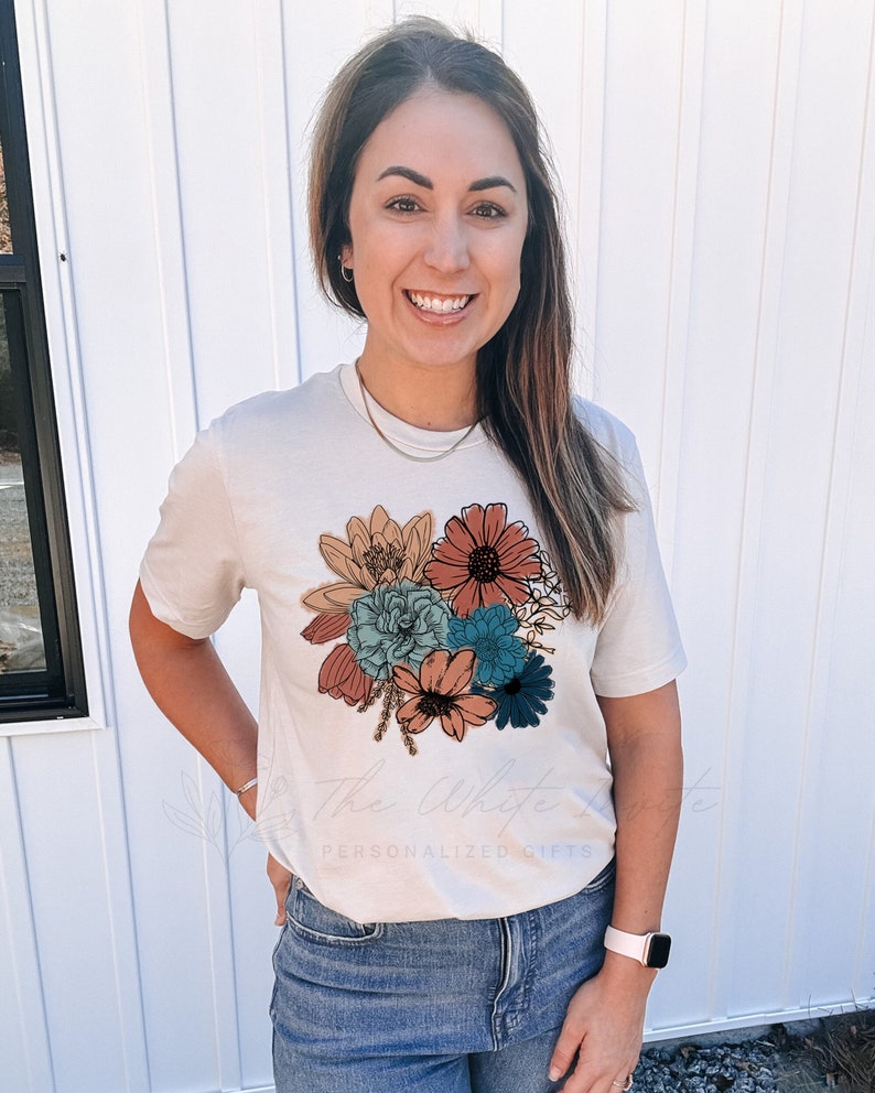 May include: A white t-shirt featuring a floral design with a variety of flowers in shades of orange, red, blue, and green. The flowers are outlined in black and have a vintage style.