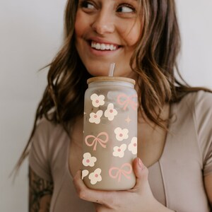 May include: A clear glass tumbler with a bamboo lid, straw, and a floral and bow design in cream and pink. The tumbler is filled with a light-colored beverage. The design includes flowers, bows, and a star.