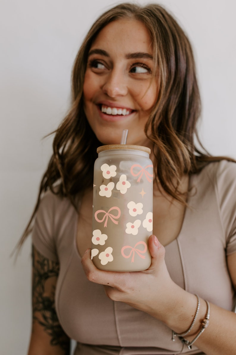 May include: A clear glass tumbler with a bamboo lid, straw, and a floral and bow design in cream and pink. The tumbler is filled with a light-colored beverage. The design includes flowers, bows, and a star.
