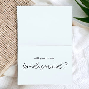 May include: A white card with black text that reads "Will you be my bridesmaid?"