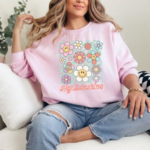 May include: Pink sweatshirt with a graphic print of colourful flowers and smiley faces. The text "Hey Sunshine" is printed below the flowers.
