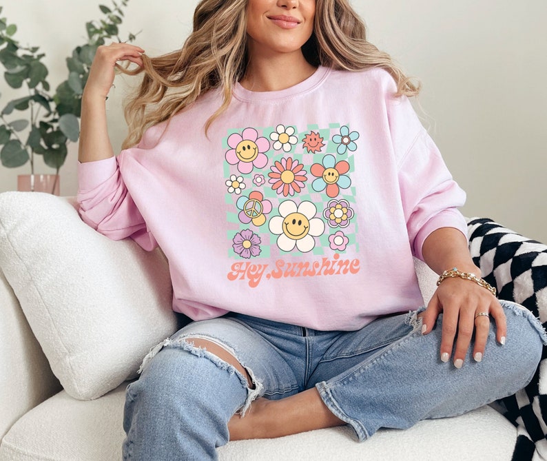 May include: Pink sweatshirt with a graphic print of colourful flowers and smiley faces. The text "Hey Sunshine" is printed below the flowers.