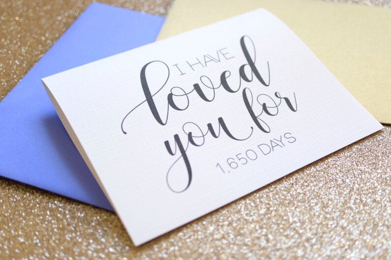May include: White card with black calligraphy that reads "I have loved you for 1,650 days". The card is sitting on a gold glitter background.
