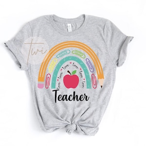 May include: Gray t-shirt with a rainbow design featuring a red apple, pencils, and paper clips. The rainbow has the words "love" written around it. The text "Teacher" is below the rainbow.
