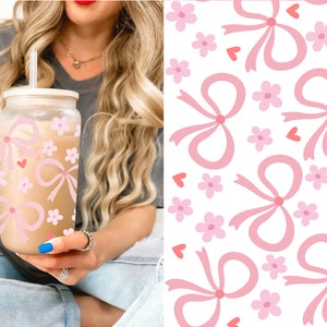May include: A glass tumbler with a bamboo lid and straw, decorated with pink bows, flowers, and hearts. The tumbler is filled with a light brown beverage. A matching pattern of bows, flowers, and hearts is on a white background.