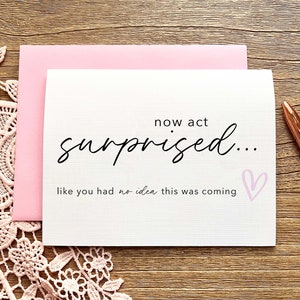 May include: A white card with pink accents and the text "now act surprised... like you had no idea this was coming" with a pink heart.