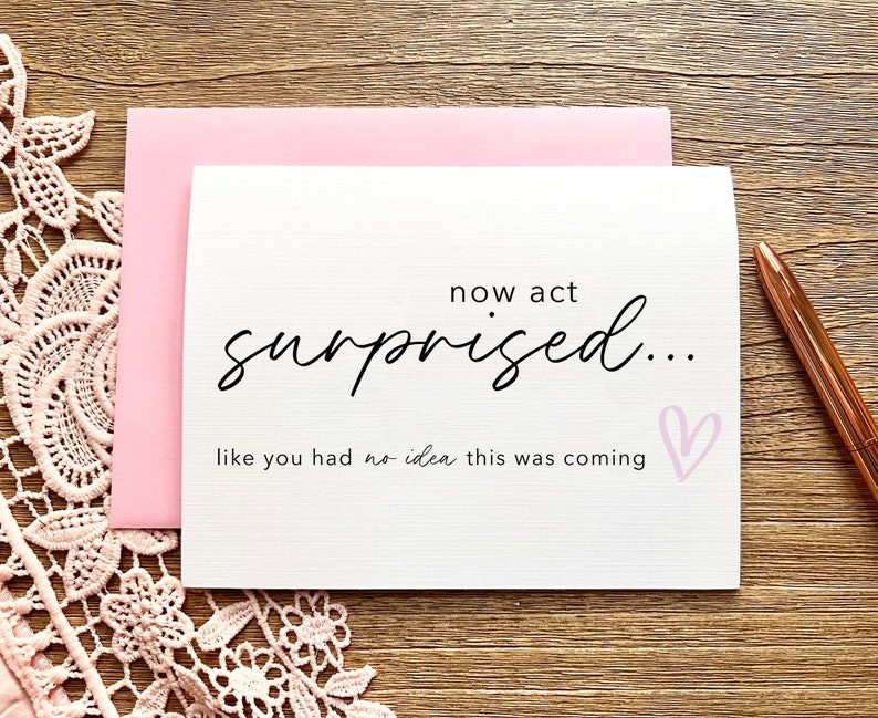 May include: A white card with pink accents and the text "now act surprised... like you had no idea this was coming" with a pink heart.