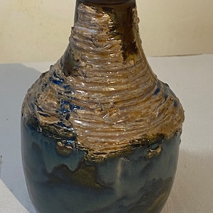 Puede incluir: Un pequeño jarrón de cerámica con un esmalte texturizado marrón y blanco. El jarrón tiene un cuerpo redondeado y un cuello estrecho. El esmalte tiene un aspecto moteado, con zonas de azul oscuro y marrón.