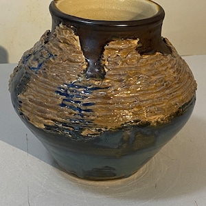 Puede incluir: Un jarrón de cerámica marrón y azul con una superficie texturizada. El jarrón tiene una boca ancha y un cuerpo redondeado.