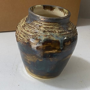 Puede incluir: Un jarrón de cerámica con un esmalte texturizado marrón y blanco. El jarrón tiene una boca ancha y un cuerpo redondeado.